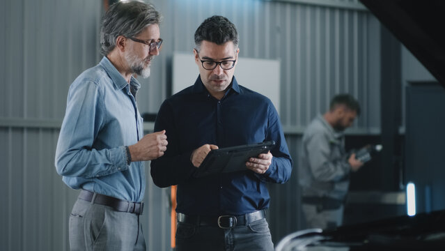 In car service manager in glasses and casual suit with a tablet talking to a man and discussing car diagnostics and repairing