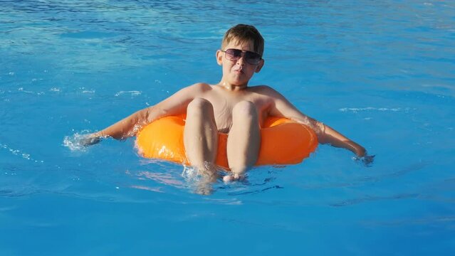 a cheerful teenager boy in sunglasses swims in the pool sitting in a swimming inflatable circle and rows with both hands like oars making a funny grimace. Swimming at sunset