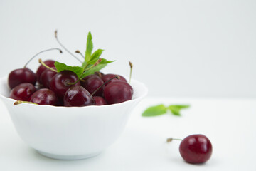 A white plate with a ripe burgundy cherry and a mint petal stands on a white background, there is a cherry and mint leaves next to it, a side view with a place for text. High quality photo
