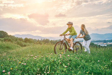 Obraz premium Mother teaching son to ride bicycle. Happy cute boy in helmet learn to riding a bike in park on green meadow in summer day at sunset time. Family weekend. 4K video Slow motion 