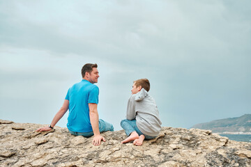 Happy family, father and son bonding, sitting on stone by the sea looking at view enjoying summer vacation. Togetherness Friendly concept
