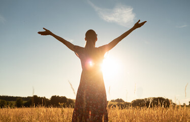 Young woman in a meadow feeing free, happy in nature with arms up facing beautiful golden sunset 