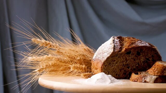 Dark bread on a wooden board with ears of wheat and flour rotates on a gray background.