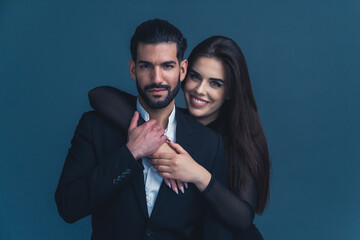 Handsome Cuban bearded guy in a black suit standing, looking at camera, and being held from the back by a beautiful long--haired caucasian girl with broad smile. Studio isolated shot. High quality