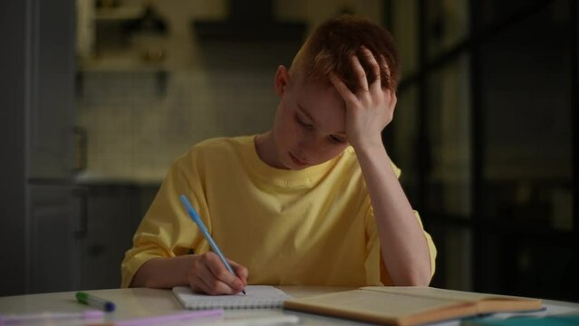 Portrait of exhausted upset schoolboy student studying alone feeling headache writing difficult assignment at home. Tired bored pupil boy worried writing in paper notebook, distance learning.