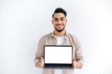 Joyful handsome Indian or Arabian young man, student or freelancer, stands on white isolated background, holding open laptop with blank white mockup screen for presentation, looking at camera, smiling