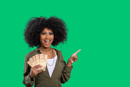 woman holding money in one hand, and point to empty space, afro woman holding brazilian money, isolated on green background