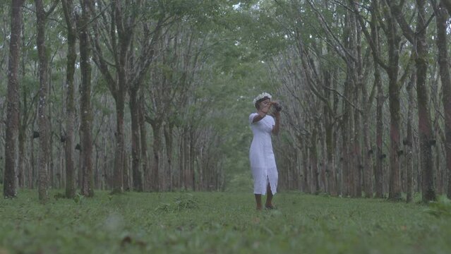 A woman in a white dress wearing a crown made of flowers stands for a picture in a rubber plantation..A tunnel of trees that looks deep and has dimensions background..4K High quality video.