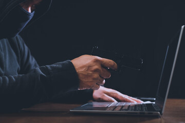 Hacker holding gun pointed at laptop symbolizes hacking and security of computer information system. © Nunkung