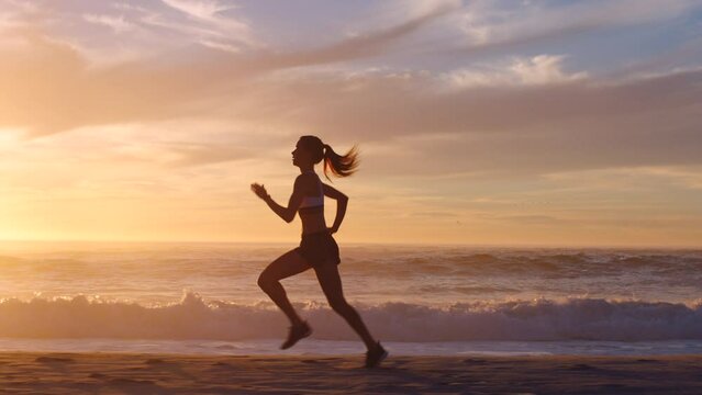 Active, fit and motivated woman running in slow motion on a beach at sunset outside. Silhouette of a determined and healthy female exercising and training outside near the ocean on a cloudy day.