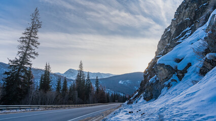 The highway runs along a steep mountain slope. Dividing lines on the asphalt, a protective barrier on the  roadside . Coniferous trees and a mountain range against the blue sky. Altai. Chuysky tract