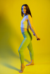 Fototapeta na wymiar Athletic girl in green leggings and a blue t-shirt does exercises for the buttocks with a resistance band. Fitness woman exercising. yellow background.