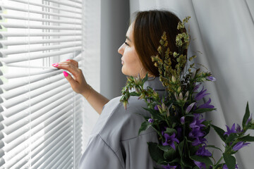 young woman in trendy gray satin trouser suit. beautiful stylish photo in the style of minimalism by the window. white studio. herbs, flowers, a bouquet behind the girl's back. The beauty