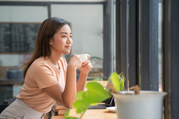 Cheerful Asian young woman drinking warm coffee or tea enjoying it while sitting in a cafe. Attractive happy Asian woman holding a cup of coffee.