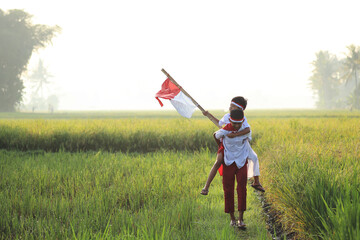  Two Indonesian elementary school students doing piggyback and walking while holding a flag during independence day in the rice field. Proud primary pupil with Indonesia flag 