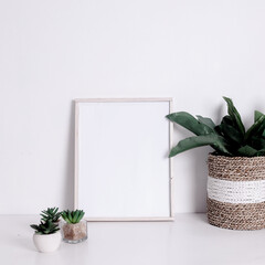 Blank wooden vertical frame and green houseplants flowers on table against white wall background. Mockup Template for your design, text.