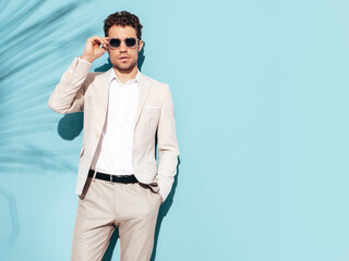 Portrait of handsome confident stylish hipster lambersexual model. Sexy modern man dressed in white elegant suit. Fashion male posing in studio near blue wall. In sunglasses