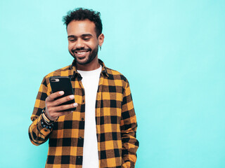 Handsome smiling hipster model. Sexy unshaven man dressed in yellow  summer shirt and jeans clothes. Fashion male posing near blue wall. Holding smartphone. Looking at cellphone screen. Using apps