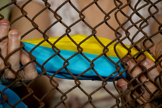 The old woman holds the flag of Ukraine with an old rusty grid in her hands, stop the war, kidnapping and theft of Ukrainians, mass slavery - Powered by Adobe