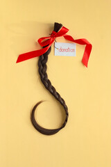Long brown braid with tag label and red ribbon on yellow background, donation of haircut, natural...