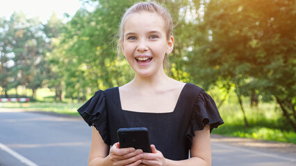Teenage girl wearing stylish black dress reads happy news on phone. Schoolgirl with braid enjoys news from friends smiling on park road, sunlight