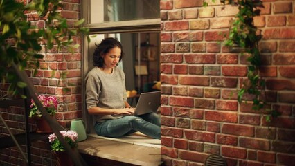 Creative Female Writer Works on a Laptop Computer while Sitting on a Windowsill of her Cozy Apartment. Professional Artist Working Remotely from Home, Listens Birds Sing. Shot Through Building Window.