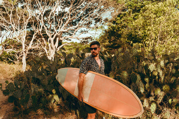 Surfer z deską surfingową, idący wybrzeżem na naturalnym tle, drzewa i kaktusy. © insomniafoto