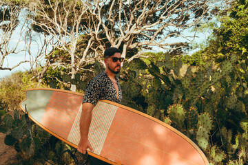 Surfer z deską surfingową, idący wybrzeżem na naturalnym tle, drzewa i kaktusy. © insomniafoto