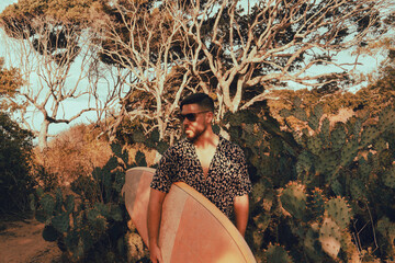 Surfer z deską surfingową, idący wybrzeżem na naturalnym tle, drzewa i kaktusy.