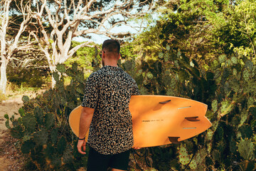 Surfer z deską surfingową, idący wybrzeżem na naturalnym tle, drzewa i kaktusy. © insomniafoto