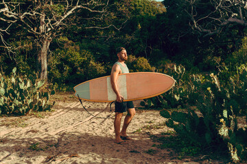Mężczyzna surfer z deską surfingową, idący wybrzeżem na tle jungle, aktywny wypoczynek.