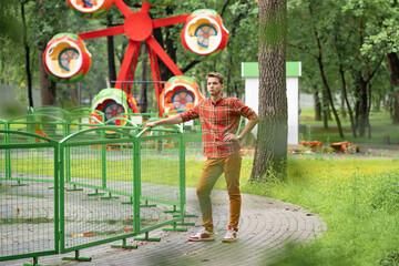 Fototapeta na wymiar a young man in a plaid shirt is standing in a park, behind him is an attraction