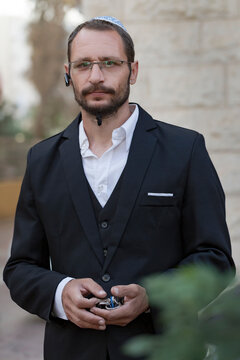 Handsome caucasian beard man with green eyes posing in the street. Orthodox jewish bearded adult guy wearing skull cap, eyeglasses, white shirt, black suit and national hat kippah  looking at camera
