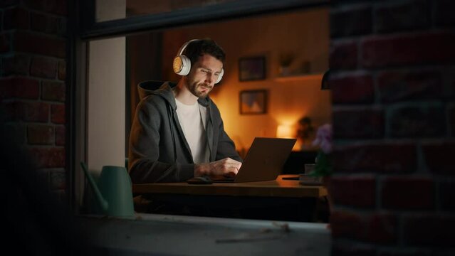 Professional Software Developer Stretches and Continues Typing on Laptop, Writing Code and Working on the Project. Remote Work at Night Time Concept. View From Outdoors into Window.