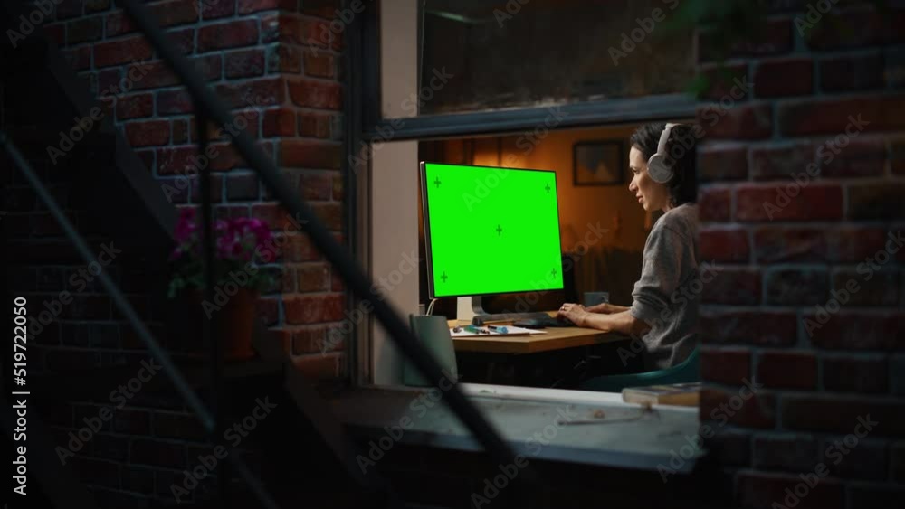 Poster Female Designer Using Desktop Computer with Green Screen Chroma Key Monitor. Remotly Working Manager Listening to Music or Podcast in Headphones, Freelancer Working at Night. Zoom Out, Window Shot.