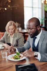 African businessman enjoying his salad sitting at the table at restaurant together with his colleague in the background
