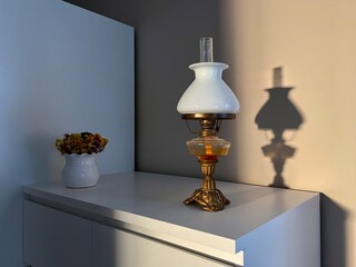 Beautiful vintage oil lamp with lamp shade made of white glass on a white cabinet. Shadow of the...
