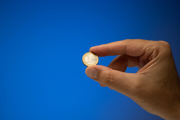 1 Euro metal coin, held in hand by a Caucasian male. Close up studio shot, isolated on blue...
