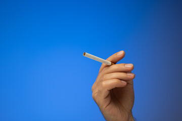 An unlit cigarette held in hand by Caucasian male hand. Close up studio shot, isolated on blue background