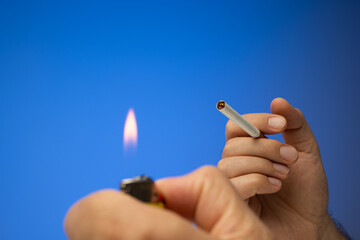 An unlit cigarette and lighter held in hand by Caucasian male hand. Close up studio shot, isolated on blue background - obrazy, fototapety, plakaty