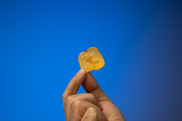 One potato chip held in hand by Caucasian male hand. Close up studio shot, isolated on blue...