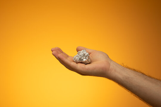 Kitchen Tin Foil Crumpled Into A Ball Held In Hand By Caucasian Male Hand. Close Up Studio Shot, Isolated On Orange Background