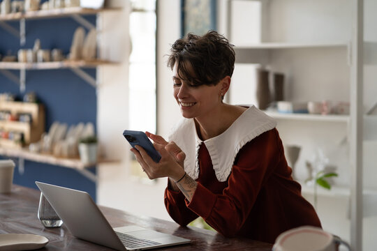 Happy successful woman small business owner chatting on mobile phone while working in pottery shop, joyful female ceramic artist newbie entrepreneur holding smartphone making first sale online - Powered by Adobe