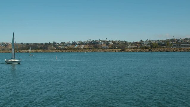 A sailboat heads out from the safe harbor of Marina Del Rey, California into the Pacific Ocean as other seacraft pass by. Seen on a summer afternoon in Los Angeles County. Static wide shot.
