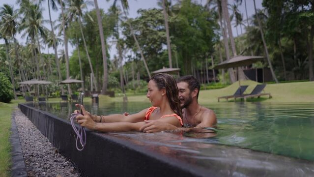 A side view of a happy young couple taking selfie photo at edge of swimming pool