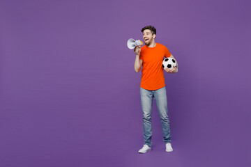 Full body young fan man he wear orange t-shirt cheer up support football sport team hold soccer ball watch tv live stream scream megaphone announces Hurry up isolated on plain dark purple background.