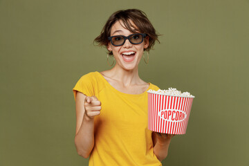 Young amazed surprised cheerful woman in 3d glasses watch movie film hold bucket of popcorn in cinema point index finger camera on you isolated on plain olive green khakibackground studio portrait.