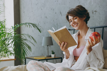Young smiling woman wear white shirt pajama she lying in bed read book study eat apple rest relax...