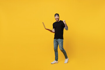 Full body young bearded tattooed man 20s he wears casual black t-shirt cap hold use work on laptop pc computer do winner gesture isolated on plain yellow wall background studio. Tattoo translate fun.