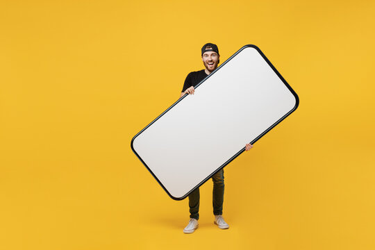 Full body happy smiling fun cool young tattooed man he 20s wears black t-shirt cap holding big huge blank screen mobile cell phone workspace mockup area copy space isolated on plain yellow background.
